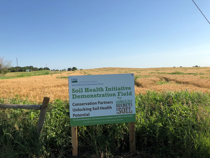 A photo shows a sign posted in front of a field with dried vegetation. The legible portions of the sign read: USDA, soil health initiative demonstration field, conservation partners unlocking soil health potential, unlock the secrets of the soil.